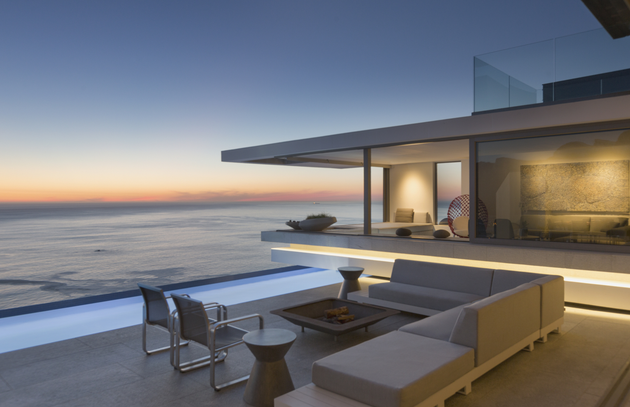 Illuminated modern luxury home with exterior patio, sofa, lap pool, and ocean view at dusk